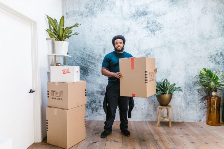 A mover carefully carries a potted plant across a snowy driveway toward a moving truck on a cold winter day.