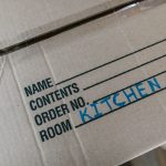 A mover in uniform carries a labeled cardboard box in a bright living room with packed items nearby.