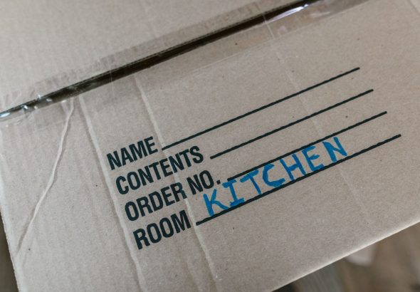 A mover in uniform carries a labeled cardboard box in a bright living room with packed items nearby.
