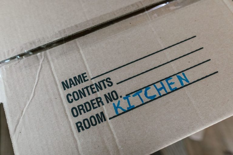 A mover in uniform carries a labeled cardboard box in a bright living room with packed items nearby.