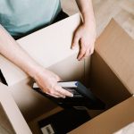 A person carefully wraps hardcover books in packing paper and places them into a small cardboard box on a living room fl