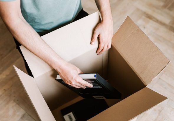 A person carefully wraps hardcover books in packing paper and places them into a small cardboard box on a living room fl