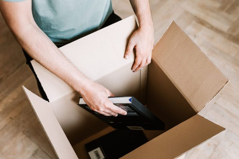 A person carefully wraps hardcover books in packing paper and places them into a small cardboard box on a living room fl