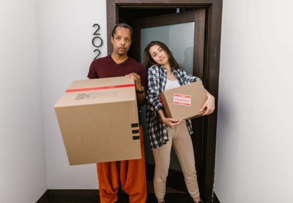 A mover carries a stack of cardboard boxes through a bright apartment hallway, preparing for moving day.