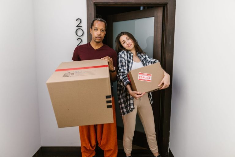 A mover carries a stack of cardboard boxes through a bright apartment hallway, preparing for moving day.