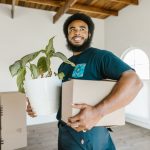 A mover carries a stack of cardboard boxes through a furnished apartment living room, with more packed boxes visible on