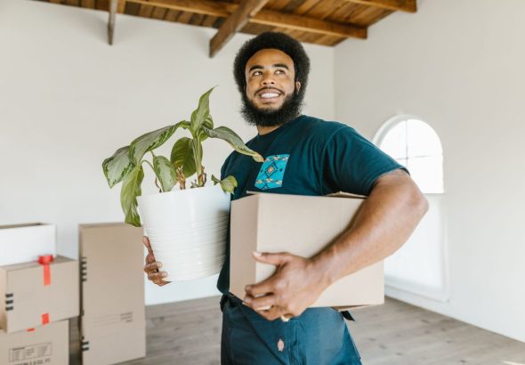 A mover carries a stack of cardboard boxes through a furnished apartment living room, with more packed boxes visible on