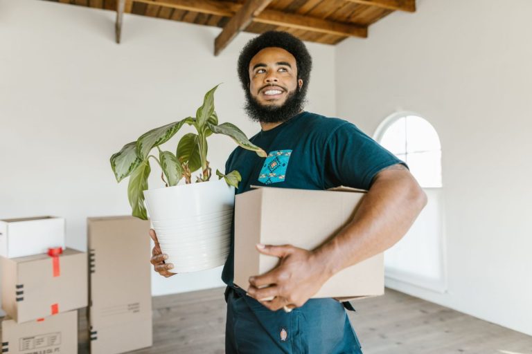 A mover carries a stack of cardboard boxes through a furnished apartment living room, with more packed boxes visible on